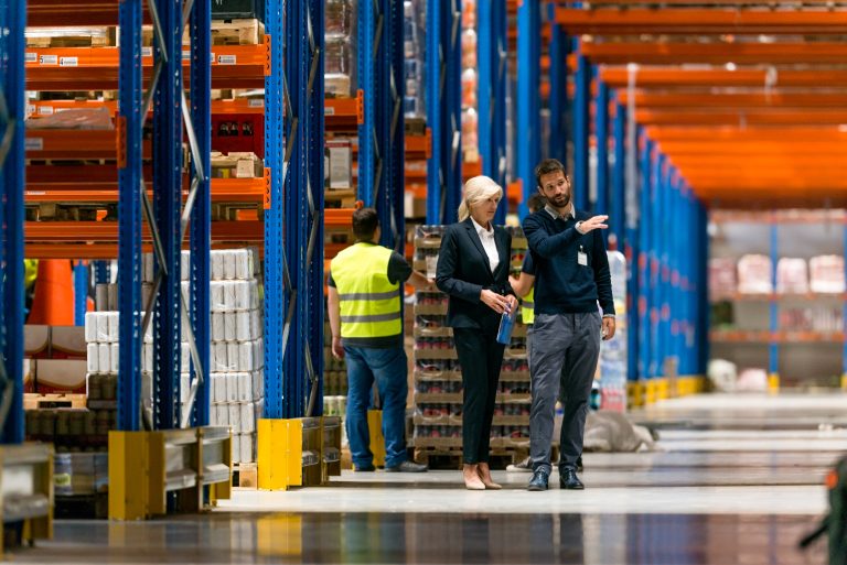 Three people coordinating work in a warehouse aisle lined with pallet racks.